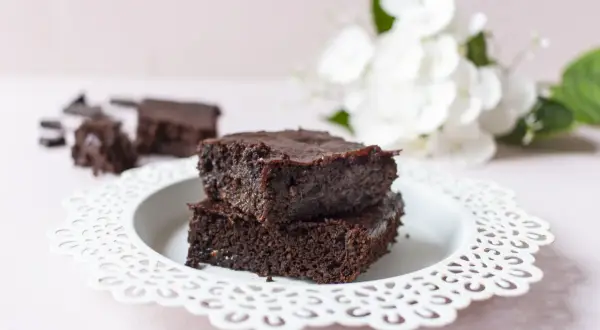 Zwei gestapelte, saftige low carb brownies auf einem weißen Teller, daneben zerbrochene Schokostücke und im Hintergrund weiße Blumen.