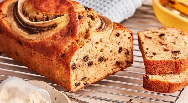 Saftiges protein bananenbrot mit Schokostückchen und Bananenscheiben, daneben eine Schüssel mit Proteinpulver.