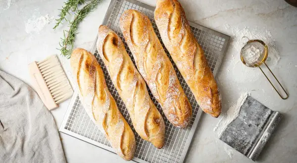 Vier frisch gebackene Baguettes auf einem Gitter, bereit für das Baguette Rezept. Daneben liegen ein Bürstchen, ein Tuch und ein Sieb mit Mehl.
