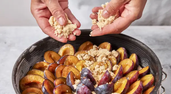 Hände streuen Streusel über saftige Pflaumen in einer Springform. Der pflaumenkuchen nimmt Gestalt an.
