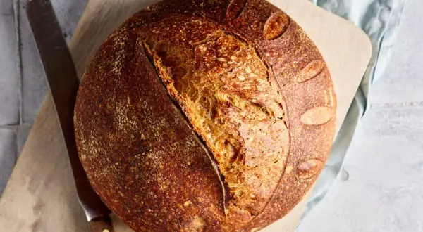 Ein rustikales Dinkel Sauerteigbrot mit knuspriger Kruste und luftiger Krume liegt auf einem Holzbrett neben einem Messer.