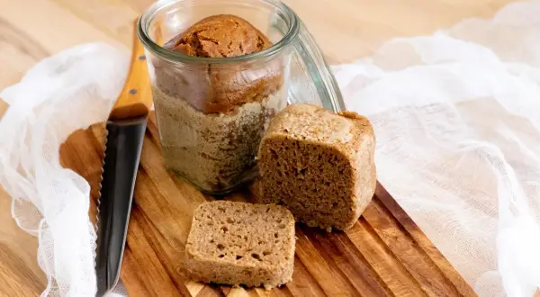 Brot im Glas Rezept: Ein Laib Brot im Glas, daneben ein Stück und ein kleines Quadrat auf einem Holzbrett. Ein Messer liegt daneben.