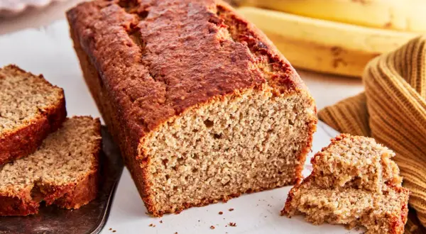Ein saftiges, goldbraunes bananenbrot glutenfrei, mit zwei Scheiben auf einem Silbertablett und reifen Bananen im Hintergrund.
