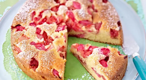 Ein saftiger Erdbeer Rhabarber Kuchen, bestäubt mit Puderzucker, liegt auf einem Teller. Ein Stück ist bereits herausgeschnitten.