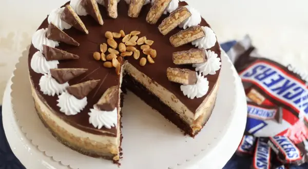 Eine köstliche Snickers Torte mit Schokoladenglasur, Erdnüssen und Sahnetuffs. Kleine Snickers-Stücke zieren den Rand.