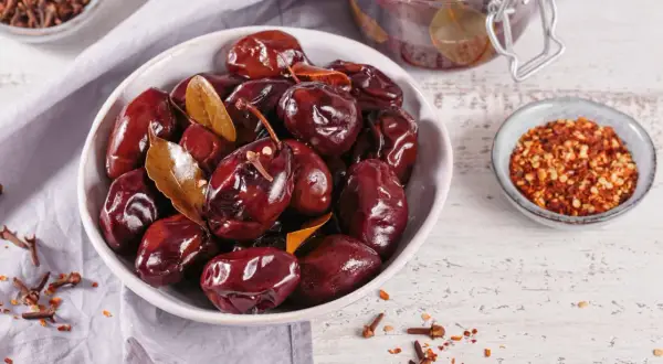 Schwarze, glänzende, fermentierte Pflaumen in einer Schüssel, bereit zum Genießen. Gewürze wie Nelken und Chiliflocken deuten darauf hin, wie man fermentierte Pflaumen selber machen kann.