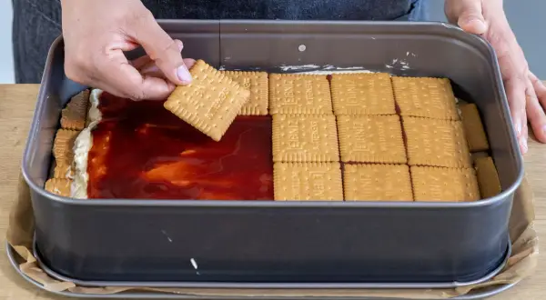 Eine Hand legt einen Butterkeks auf eine Schicht Marmelade und Creme in einer Backform. Der Butterkeks Kuchen wird geschichtet.