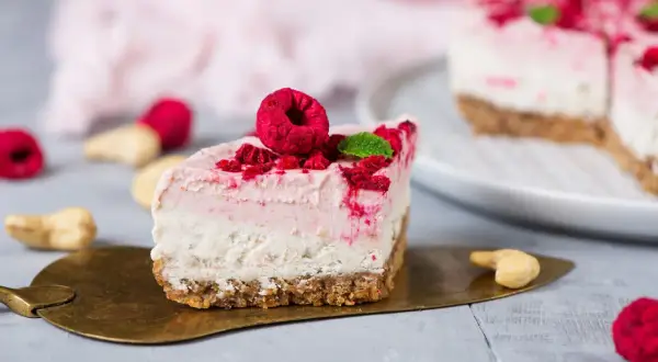 Ein Stück Himbeer Cheesecake mit frischen Himbeeren und Minze auf einem goldenen Tortenheber.