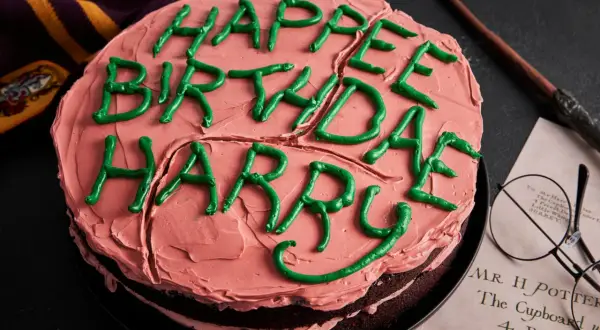 Ein Harry Potter Kuchen mit rosa Zuckerguss und grüner Schrift "Happy Birthday Harry". Daneben liegen eine Brille und ein Zauberstab.