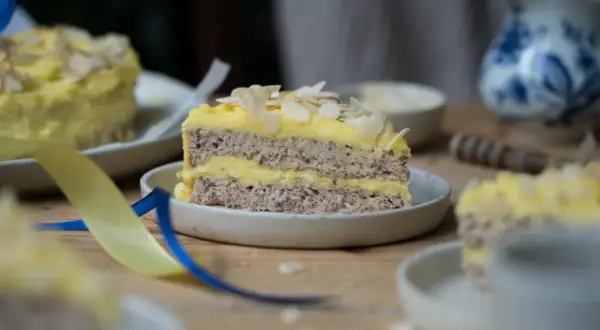 Ein Stück schwedische Mandeltorte mit gelber Creme und Mandelblättchen, dekoriert mit blauen und gelben Bändern.