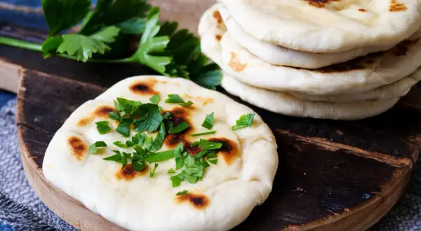 Frisch gebackenes naan brot, bestreut mit Petersilie, liegt auf einem Holzbrett neben einem Stapel weiterer Fladenbrote.