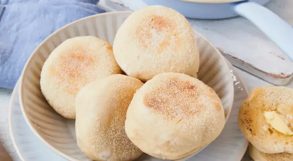 Ein Teller mit vier goldbraunen englischen Muffins, daneben ein halber, mit Butter bestrichener Muffin. Ein Spiegelei ist im Hintergrund zu sehen.