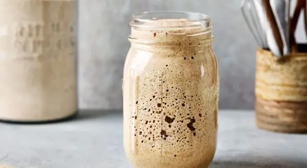 Ein Glas mit sprudelndem Sauerteig, bereit zum Backen. Im Hintergrund ein weiteres Glas mit Mehl und Küchenutensilien.
