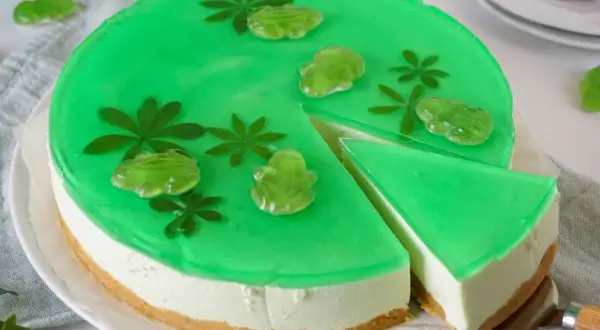 Grüne Philadelphia Torte Waldmeister mit Gummifröschen und Blättern verziert. Ein Stück wird gerade herausgeschnitten.