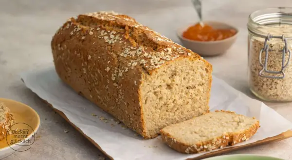 Frisch gebackenes Haferbrot, bestreut mit Haferflocken. Ein Glas Haferflocken und Marmelade daneben deuten auf ein leckeres Haferbrot Rezept hin.