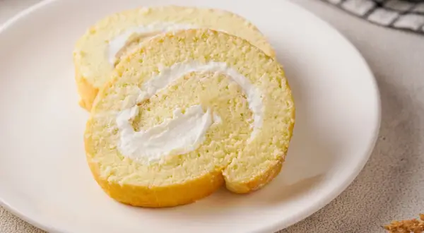 Zwei Scheiben einer saftigen Zitronenrolle mit cremiger Füllung auf einem weißen Teller. Ein köstliches Zitronenrolle Rezept zum Nachbacken.