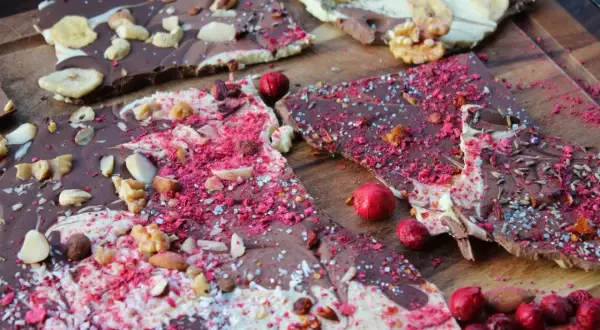 Bruchschokolade selber machen: Bunte Schokoladenstücke mit Nüssen, Beeren und Bananenchips auf Holzbrett.