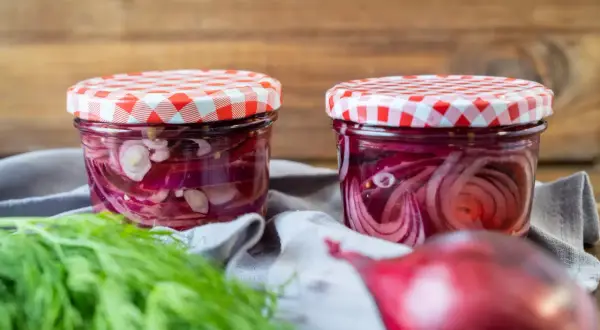 Zwei Gläser mit eingelegten roten Zwiebeln, bereit für Salate oder Sandwiches. Frischer Dill liegt daneben.