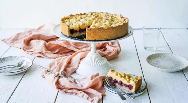 Ein saftiger Kirschkuchen mit Pudding und Streuseln auf einem Kuchenständer. Ein Stück ist bereits auf einem Teller angerichtet.