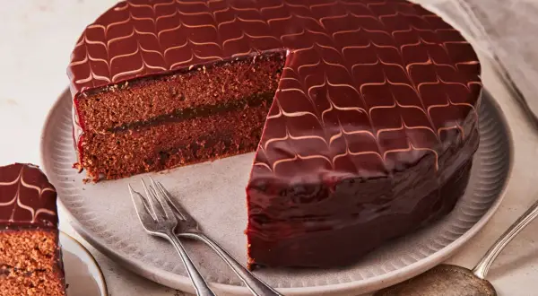 Eine glänzende Sacher Torte mit Schokoladenglasur und einem Stück Marmeladenfüllung. Gabeln liegen bereit.