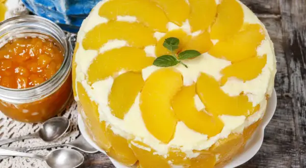 Eine köstliche Pfirsich Maracuja Torte, dekoriert mit frischen Pfirsichspalten und Minze. Daneben ein Glas Pfirsichmarmelade.