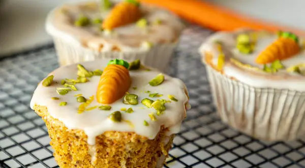 Drei leckere Karotten Muffins mit weißer Glasur, gehackten Pistazien und kleinen Marzipan-Karotten auf einem Gitter.