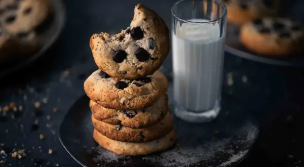 Ein Stapel Schokoladenkekse, eines davon angeknabbert, neben einem Glas Milch. Perfekt für ein einfaches Cookies Rezept.