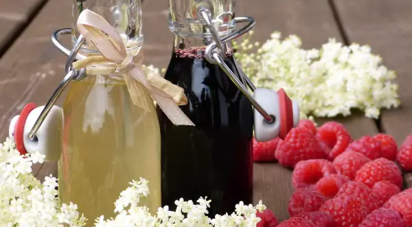 Zwei Flaschen mit selbstgemachtem Sirup, umgeben von Holunderblüten und Himbeeren. Perfekt zum Sirup einkochen.