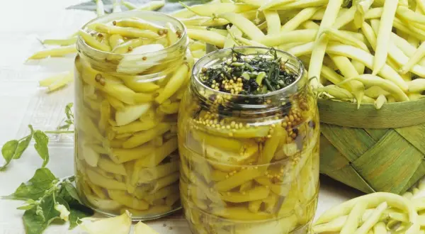 Zwei Gläser mit eingelegten Buschbohnen, daneben frische Bohnen und Knoblauch.