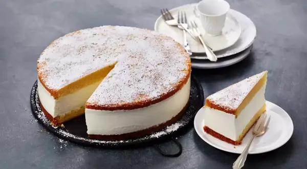 Eine köstliche Quark Sahne Torte, bestäubt mit Puderzucker, wartet auf Genießer. Ein Stück ist bereits auf einem Teller angerichtet.