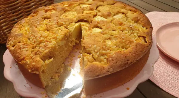 Ein saftiger apfelkuchen ohne zucker, bestreut mit Nüssen, wird auf einem rosa Teller präsentiert. Daneben liegt ein roter Apfel.