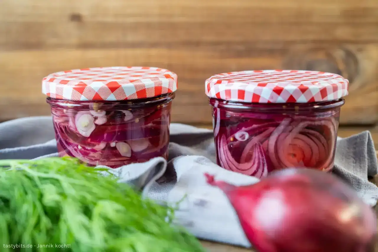 Zwei Gläser mit eingelegten roten Zwiebeln, bereit für Salate oder Sandwiches. Frischer Dill liegt daneben.