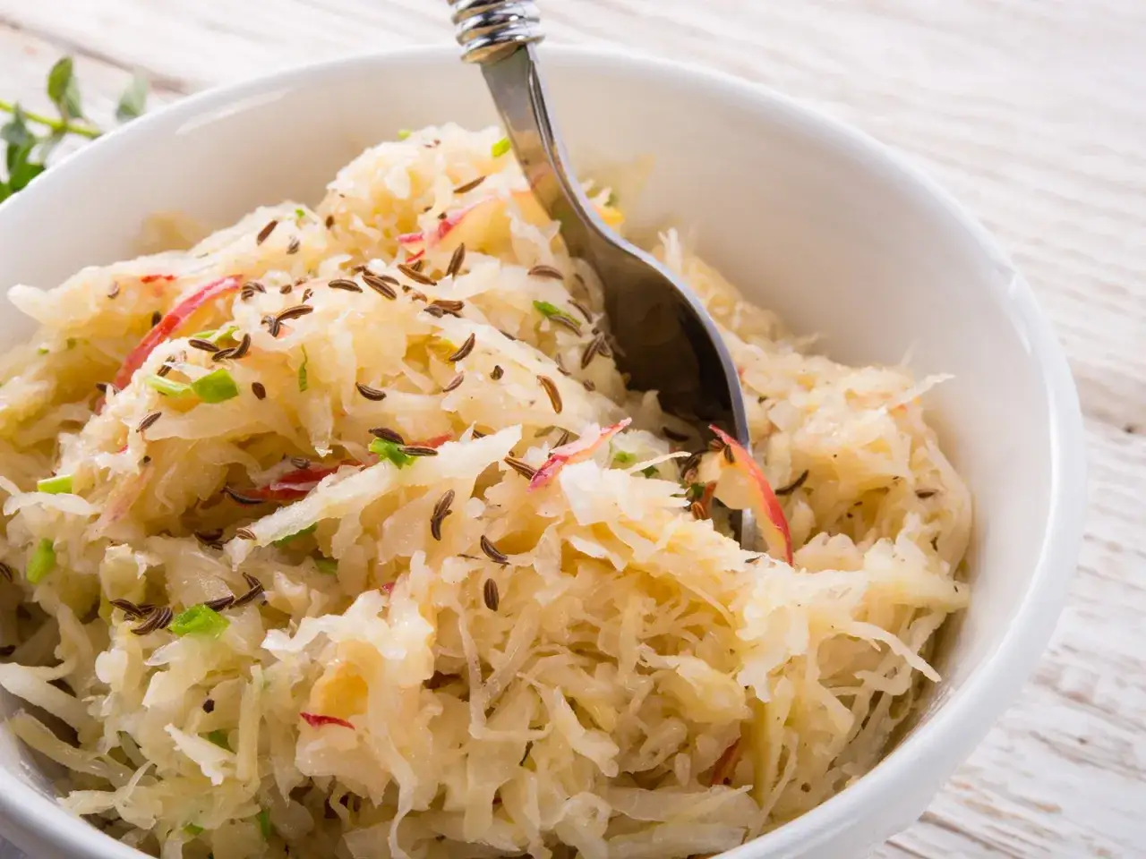 Schüssel mit Sauerkraut, Kümmel und Apfelstücken. Ein Löffel ragt hinein. Perfekt für Ihr Sauerkraut Rezept.