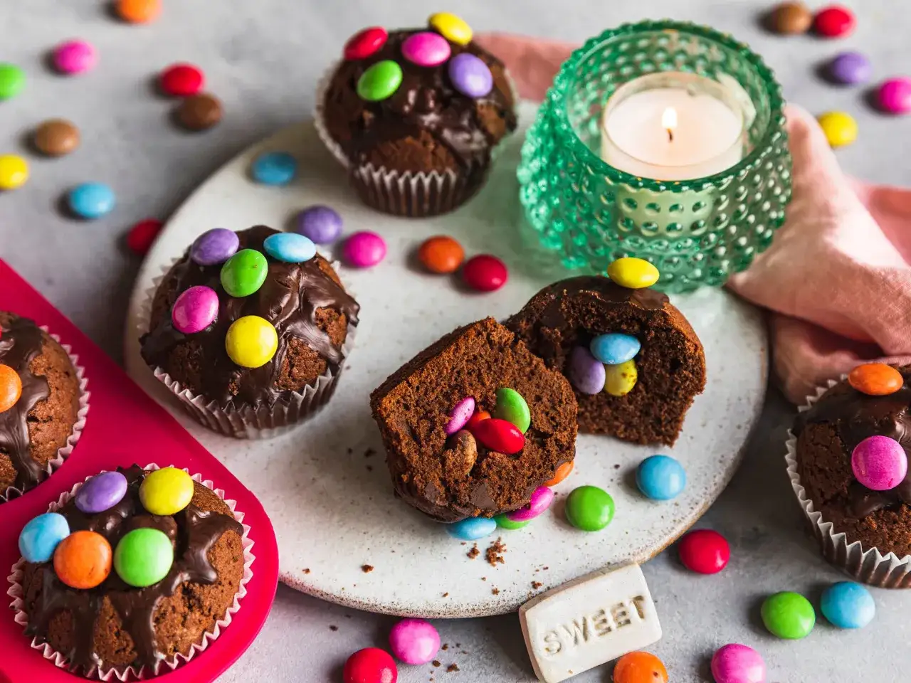 Schokoladenmuffins mit bunten Smarties, perfekt für einen Kindergeburtstag. Ein Teelicht in einem grünen Kerzenhalter sorgt für festliche Stimmung.
