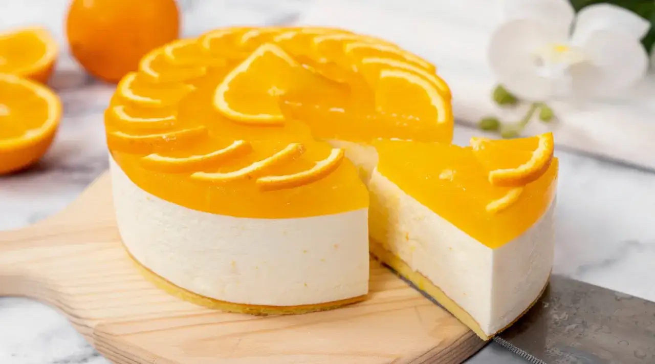 Eine leuchtend orangefarbene Aperol Torte mit frischen Orangenscheiben, auf einem Holzbrett, daneben eine Scheibe und Orangen.