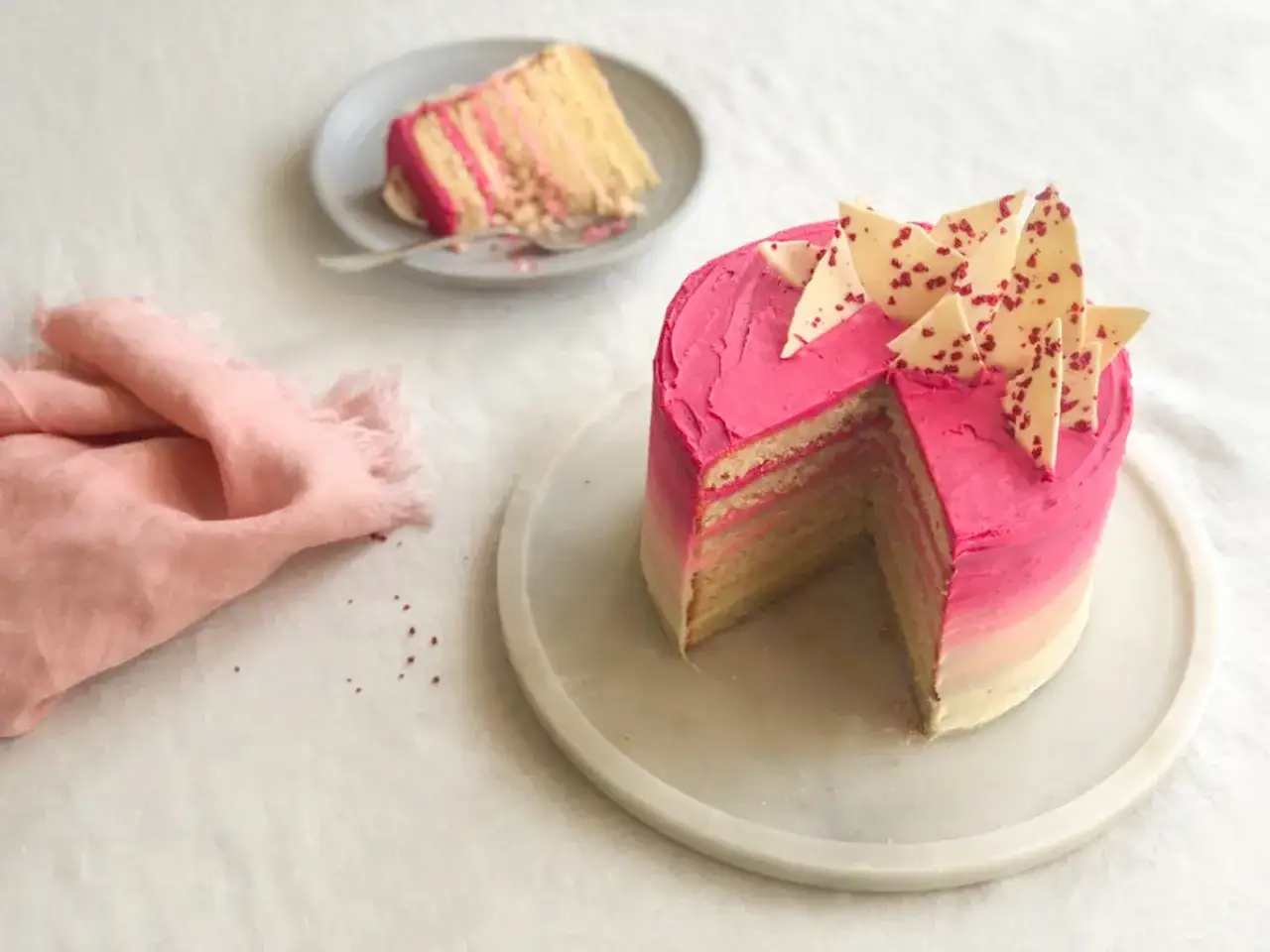 Eine mehrschichtige Buttercreme Torte mit pinkem Ombre-Frosting und wei&szlig;en Schokoladenst&uuml;cken. Ein St&uuml;ck ist bereits auf einem Teller.