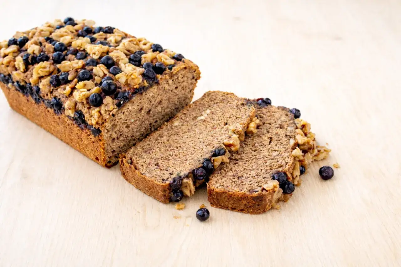 Ein saftiges Bananenbrot, glutenfrei, mit Blaubeeren und Walnüssen belegt. Drei Scheiben liegen daneben.