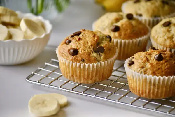 Frisch gebackene schoko bananen muffins mit Schokostückchen auf einem Kuchengitter. Daneben eine Schüssel mit Bananenscheiben.