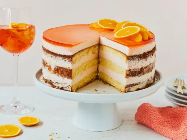 Eine Aperol Torte mit fruchtiger Geleehaube und Orangenscheiben, daneben ein Aperol Spritz.