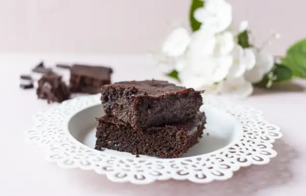 Zwei gestapelte, saftige low carb brownies auf einem weißen Teller, daneben zerbrochene Schokostücke und im Hintergrund weiße Blumen.