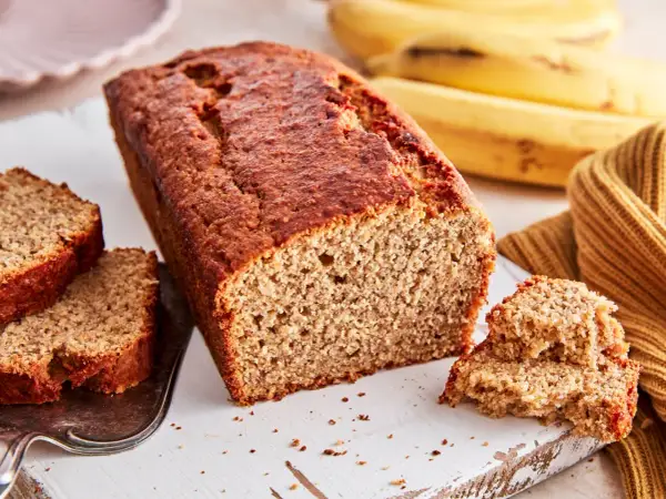Ein saftiges, goldbraunes bananenbrot glutenfrei, mit zwei Scheiben auf einem Silbertablett und reifen Bananen im Hintergrund.