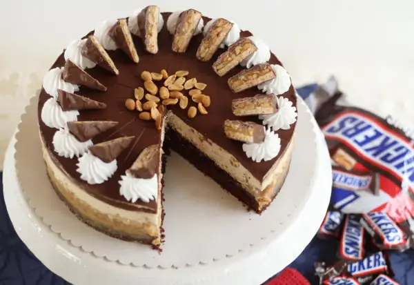 Eine köstliche Snickers Torte mit Schokoladenglasur, Erdnüssen und Sahnetuffs. Kleine Snickers-Stücke zieren den Rand.