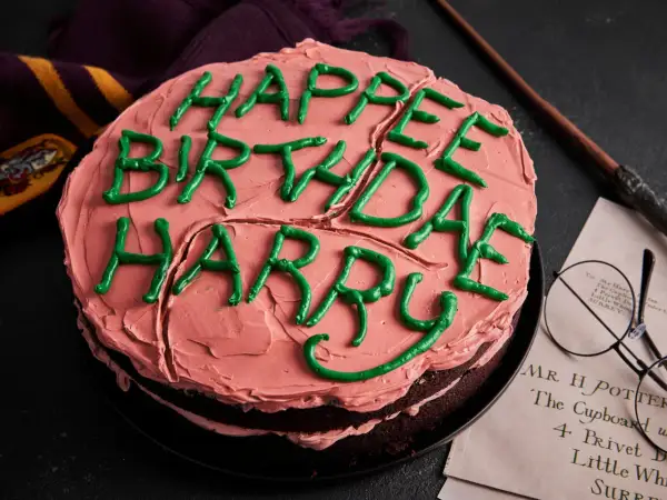 Ein Harry Potter Kuchen mit rosa Zuckerguss und grüner Schrift "Happy Birthday Harry". Daneben liegen eine Brille und ein Zauberstab.