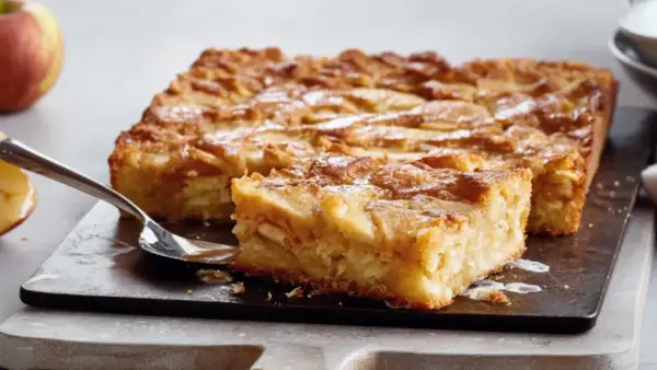 Saftige schwedische Apfeltorte, mit Äpfeln belegt und mit Puderzucker bestäubt, auf einem dunklen Brett mit einer Gabel.