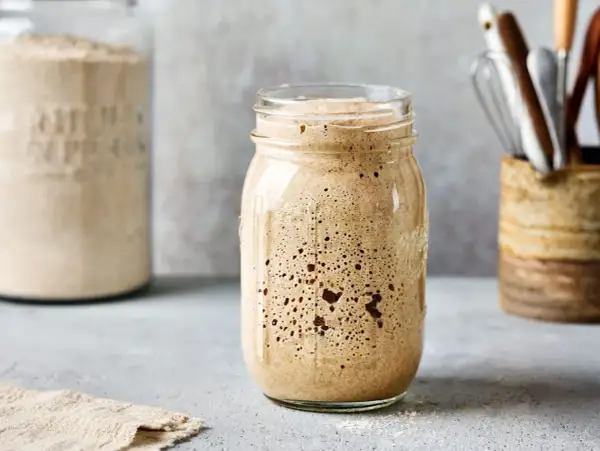 Ein Glas mit sprudelndem Sauerteig, bereit zum Backen. Im Hintergrund ein weiteres Glas mit Mehl und Küchenutensilien.