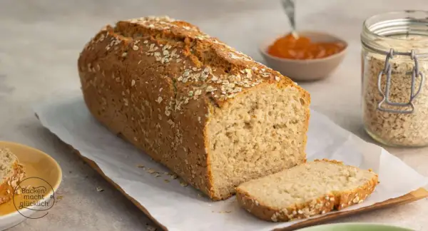 Frisch gebackenes Haferbrot, bestreut mit Haferflocken. Ein Glas Haferflocken und Marmelade daneben deuten auf ein leckeres Haferbrot Rezept hin.