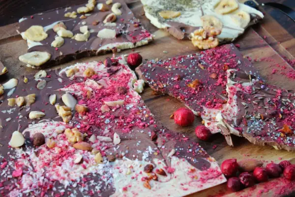 Bruchschokolade selber machen: Bunte Schokoladenstücke mit Nüssen, Beeren und Bananenchips auf Holzbrett.
