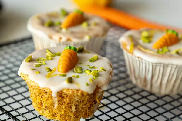 Drei leckere Karotten Muffins mit weißer Glasur, gehackten Pistazien und kleinen Marzipan-Karotten auf einem Gitter.