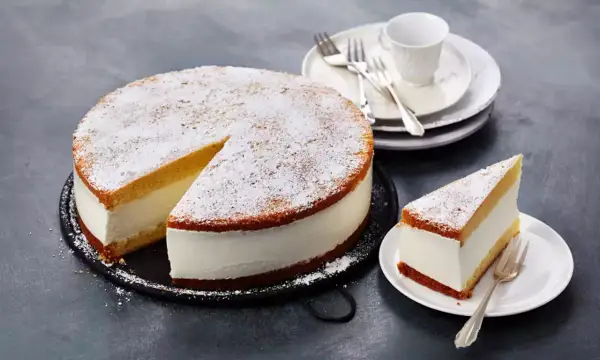 Eine köstliche Quark Sahne Torte, bestäubt mit Puderzucker, wartet auf Genießer. Ein Stück ist bereits auf einem Teller angerichtet.