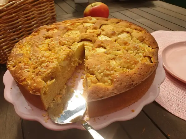 Ein saftiger apfelkuchen ohne zucker, bestreut mit Nüssen, wird auf einem rosa Teller präsentiert. Daneben liegt ein roter Apfel.